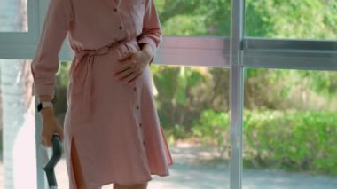 Bir stok videosunda pembe elbise giyen hamile bir kadın kablosuz elektrikli süpürge kullanarak yerleri zarafetle temizliyor. Kamera onu panoramik bir pencerenin yanında dururken yakalar.
