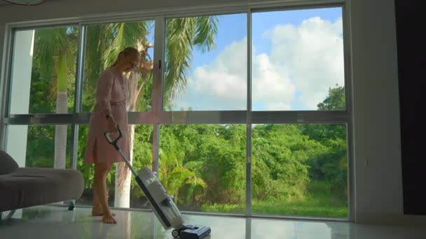 一段视频展示了一名孕妇穿着粉色连衣裙 用无绳电动拖把吸尘器优雅地清扫地板的情景 摄像机捕捉到她站在全景窗前 — 图库视频影像