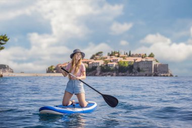 Genç kadınlar, Karadağ 'ın St. Stefan adası yakınlarındaki mavi denizde ayakta kürek çekerken eğleniyorlar. Çeviri:.