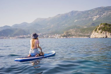 Genç kadınlar, Karadağ 'daki Milocer Park' ın arka planına karşı St. Stefan adası yakınlarındaki mavi su denizinde ayakta kürek çekerken eğleniyorlar. Çeviri:.