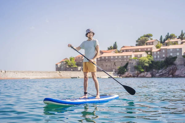 在黑山圣斯特凡岛附近的蓝色海面上 有一个年轻的男人站着玩得很开心 Sup — 图库照片