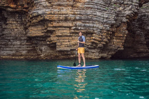 在黑山的岩石中间 有一个年轻人在蓝色的海水中打起精神来 Sup — 图库照片