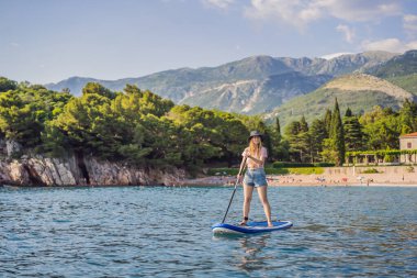 Genç kadınlar, Karadağ 'daki Milocer Park' ın arka planına karşı St. Stefan adası yakınlarındaki mavi su denizinde ayakta kürek çekerken eğleniyorlar. Çeviri:.