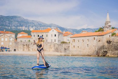 Genç kadınlar, Karadağ 'ın mavi sularında ayakta kürek çekerken eğleniyorlar. Eski Budva Kasabası 'nın arka planına karşı. Çeviri:.