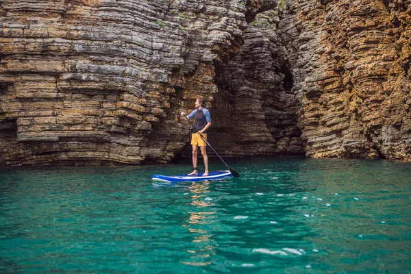 在黑山的岩石中间 有一个年轻人在蓝色的海水中打起精神来 Sup — 图库照片