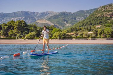 Genç erkekler, Karadağ 'ın St. Stefan adası yakınlarındaki mavi denizde ayakta kürek çekerken eğleniyorlar. Çeviri:.