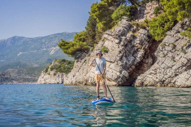 Genç erkekler, Karadağ 'daki Milocer Park' ın arka planına karşı St. Stefan adası yakınlarındaki mavi su denizinde ayakta kürek çekerek eğleniyorlar. Çeviri:.