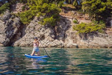 Genç kadınlar, Karadağ 'daki Milocer Park' ın arka planına karşı St. Stefan adası yakınlarındaki mavi su denizinde ayakta kürek çekerken eğleniyorlar. Çeviri:.