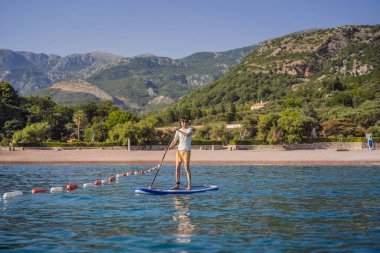 Genç erkekler, Karadağ 'ın St. Stefan adası yakınlarındaki mavi denizde ayakta kürek çekerken eğleniyorlar. Çeviri:.