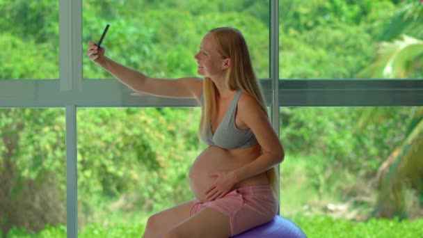 スマートフォンを持った妊婦さんが大きな窓の前に位置し 緑豊かな熱帯の背景を見せてくれます 彼女は自撮りをしている その視覚的な表現は — ストック動画