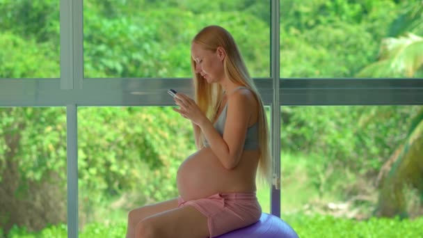 一位孕妇优雅地坐在一个健康的球上 放置在一个巨大的橱窗前 展示着茂盛的绿色热带背景 她手里拿着一部电话 从事着各种各样的活动 — 图库视频影像