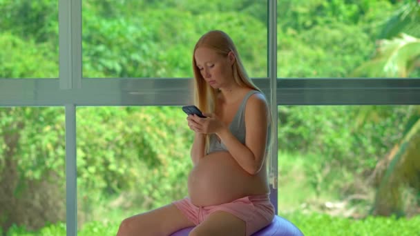 妊娠中の女性は 緑豊かな熱帯の背景を示す大きな窓の前に配置されたフィットボールの上に優雅に座っています 彼女の手に電話をかけ 彼女はさまざまな活動に従事しています — ストック動画
