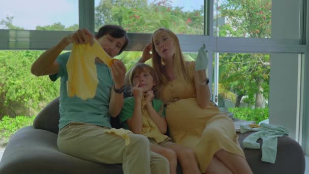 居心地の良いソファに座っている父親 妊婦の家族を示す心温まるストックビデオ すぐに到着する小さな服のために愛らしい小さな服を楽しくブラウズしてください — ストック動画