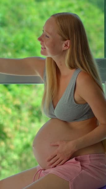 バーティカルビデオ ストーリーフォーマット スマートフォンを持つ妊婦は 緑豊かな熱帯の背景を示す大きな窓の前に位置するフィットボールに座っています 彼女は自分撮りをしている — ストック動画