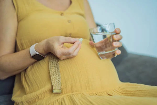 Prenatal Vitamins. Portrait Of Beautiful Smiling Pregnant Woman Holding Supplement and a glass of water, Taking Supplements For Healthy Pregnancy While Sitting On Couch At Home, Free Space.