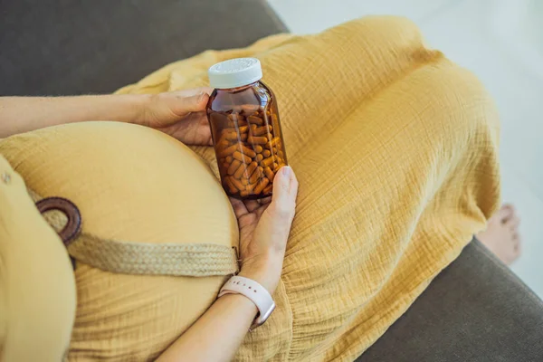 Prenatal Vitamins. Portrait Of Beautiful Smiling Pregnant Woman Holding Transparent Glass Jar With Pills, Taking Supplements For Healthy Pregnancy While Sitting On Couch At Home, Free Space.