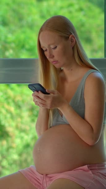 バーティカルビデオ ストーリーフォーマット 妊娠中の女性は 緑豊かな熱帯の背景を示す大きな窓の前に配置されたフィットボールの上に優雅に座っています 彼女の手に電話をかけると 彼女は — ストック動画