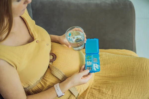 Prenatal Vitamins. Portrait Of Beautiful Smiling Pregnant Woman Holding Pill Box and a glass of water, Taking Supplements For Healthy Pregnancy While Sitting On Couch At Home, Free Space.