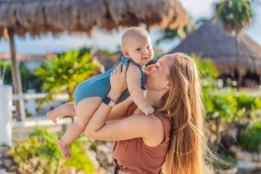 Tropikal palmiyeler ve sazdan çatılar arasında, sevgi dolu bir anne bebeğini kucaklar, sıcaklığını ve sevgisini açık havada paylaşır..