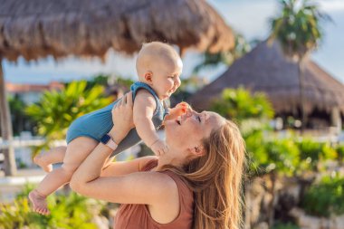 Tropikal palmiyeler ve sazdan çatılar arasında, sevgi dolu bir anne bebeğini kucaklar, sıcaklığını ve sevgisini açık havada paylaşır..