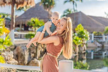 Tropikal palmiyeler ve sazdan çatılar arasında, sevgi dolu bir anne bebeğini kucaklar, sıcaklığını ve sevgisini açık havada paylaşır..