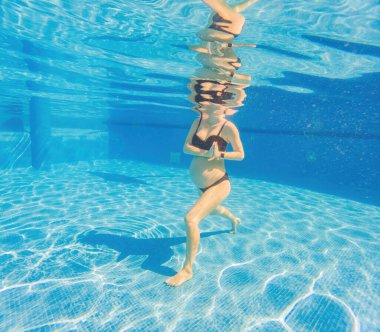 Su sporunu kucaklayan hamile bir kadın, su altı aerobiğinde güç ve dinginlik gösteriyor. Havuzda dingin ve güçlü bir görüntü yaratıyor..