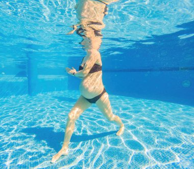 Su sporunu kucaklayan hamile bir kadın, su altı aerobiğinde güç ve dinginlik gösteriyor. Havuzda dingin ve güçlü bir görüntü yaratıyor..