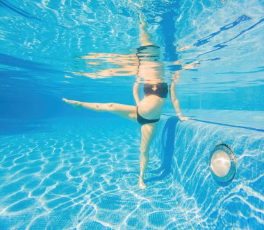 Su sporunu kucaklayan hamile bir kadın, su altı aerobiğinde güç ve dinginlik gösteriyor. Havuzda dingin ve güçlü bir görüntü yaratıyor..