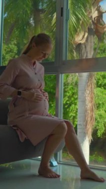 Dikey video. Kablosuz elektrikli süpürge kullanarak zarif bir şekilde yerleri temizleyen pembe elbiseli hamile bir kadın. Kamera onu panoramik pencerenin yanında görüntüler.