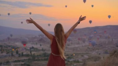 Kapadokya, Türkiye 'de genç bir kadın sıcak hava balonlarına bakıyor. İyi yolculuklar hindi konsepti. Dağın tepesindeki kadın harika manzaranın tadını çıkarıyor.