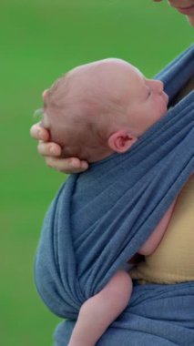 Dikey video. Yeni doğmuş küçük bir çocuk yumuşak bir bebek askısının içine sokulur. Bebekliğin narin güzelliğini yakalıyor bebek huzur içinde dinlenirken, bakıcılarının kalbine yakın bir yerde.
