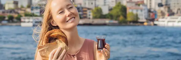BANNER, LONG FORMAT Woman in Istanbul having breakfast with Simit and a glass of Turkish tea. Glass of Turkish tea and bagel Simit against golden horn bay and the Galata Tower in Istanbul, Turkey