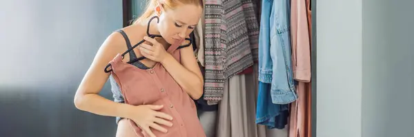 BANNER, LONG FORMAT A pregnant woman has nothing to wear. A pregnant woman stands in front of a closet with clothes and does not know what to wear because the clothes do not fit on her.