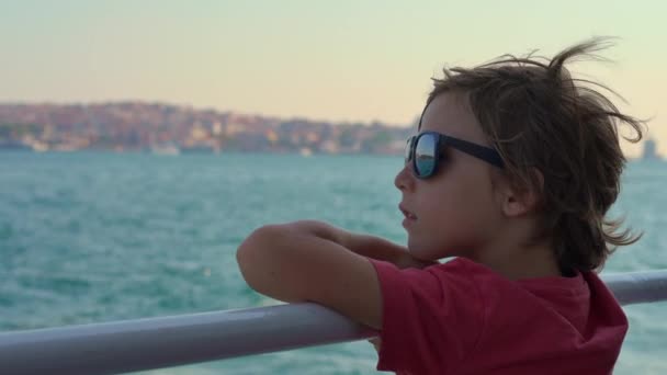 作为一个戴着太阳镜的男孩 他的目光被横扫伊斯坦布尔的景色所吸引 当渡船冲破波浪时 城市生活的背景在他身后展开 慢动作视频 — 图库视频影像