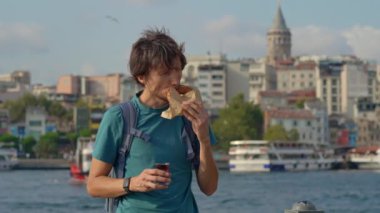 Genç bir gezgin, sert bir simit ısırığından zevk alır ve martıların dalgalandığı bir iskelede Türk çayını yudumlar ve arka planda beliren ikonik Galata Kulesi