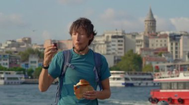 Genç bir gezgin, sert bir simit ısırığından zevk alır ve martıların dalgalandığı bir iskelede Türk çayını yudumlar ve arka planda beliren ikonik Galata Kulesi