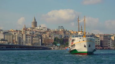 İstanbullar 'ın ikonik Galata Kulesi' nin çarpıcı manzarası şehrin siluetinin üzerinde görkemli bir şekilde yükseliyor. Ön planda bir feribot var. Yavaş çekim videosu