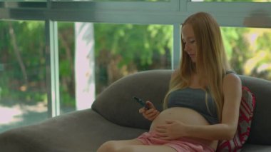 Genç hamile bir kadın pencerenin kenarındaki koltukta rahat bir şekilde oturur, akıllı telefonuna dalmış, doğal ışığın yumuşak parıltısıyla güneşlenmiştir.