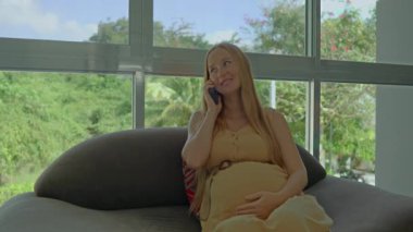 Genç hamile bir kadın pencerenin kenarındaki koltukta rahat bir şekilde oturur, akıllı telefonuna dalmış, doğal ışığın yumuşak parıltısıyla güneşlenmiştir.