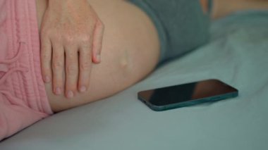 Hamile bir kadının karnının yakın plan fotoğrafı ve yanında akıllı telefon var. Telefonun ekranı kolayca değiştirilebilir ve bu video beklenen ürün ve hizmetlerin tanıtımı için mükemmel hale getirilebilir.