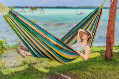 Bacalar Gölü 'nün turkuaz sularının arka planında hamakta oturan dişi turist. Aile bağı, yaz tatili ve Quintana Roo 'da açık hava eğlencesi, Meksika konsepti..