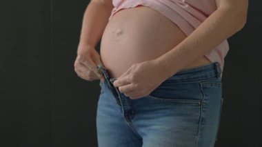 Hamile bir kadın, büyüyen karnını kotuna sığdırmak için çabalıyor ve hamileliği boyunca vücut değişikliklerinin zorluklarını vurguluyor. Bu uygun ve esprili an rahat olma ihtiyacını yansıtıyor.