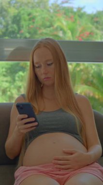 Düşünceli genç hamile bir kadın pencerenin yanındaki koltukta oturur. Yüzündeki ifade, akıllı telefon kullanırken ciddiyet ve paniklemenin bir karışımıdır..