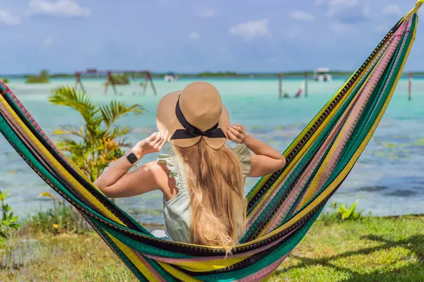 Bacalar Gölü 'nün turkuaz sularının arka planında hamakta oturan dişi turist. Aile bağı, yaz tatili ve Quintana Roo 'da açık hava eğlencesi, Meksika konsepti..