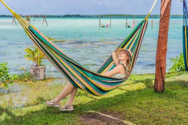 Bacalar Gölü 'nün turkuaz sularının arka planında hamakta oturan dişi turist. Aile bağı, yaz tatili ve Quintana Roo 'da açık hava eğlencesi, Meksika konsepti..