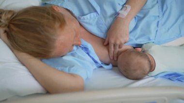 Genç bir kadın klinik bir yatakta uzanıp yeni doğmuş çocuğunu ilk kez emziriyor. Hassas ve samimi bir bağ kurma, beslenme ve hastane ortamında anneliğin başlangıcı.