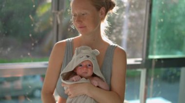 Genç bir anne sıcak bir yaz gününde güneşli bir pencerenin önünde dururken yeni doğmuş bebeğini nazikçe havluya sarar. Huzurlu ve hassas bir an. Sevgi, özen ve doğal ışıkta kaynaşma..