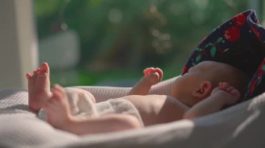 Yeni doğmuş bir bebek, güneş ışığının açık olduğu bir pencerenin yakınındaki bebek kozasında dinlenir ve sağlıklı dozda D vitamini için doğal güneş ışığını emer. Yeni doğanlar için güneşlenmenin önemini vurgulayan huzurlu bir an.