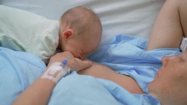 Genç bir kadın klinik bir yatakta uzanıp yeni doğmuş çocuğunu ilk kez emziriyor. Hassas ve samimi bir bağ kurma, beslenme ve hastane ortamında anneliğin başlangıcı.
