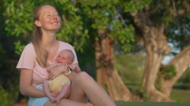 Genç bir kadın yemyeşil bir çimenlikte gülerek oturuyor, yeni doğmuş bebeğini elinde tutuyor. Doğayla çevrili neşeli ve hassas bir bağ, aşk ve mutluluk anı.